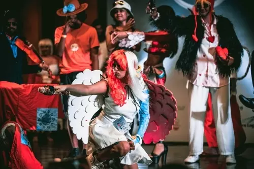 A cosplayer dressed in a colorful costume with large wings poses dramatically on stage. Other costumed participants stand around her, some wearing hats and unique outfits. The setting appears to be a lively cosplay event or competition.