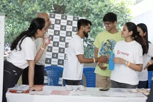 A group of people interact at a booth during an outdoor event. Two individuals behind the table engage with visitors, while others smile and talk nearby. A logo-covered backdrop and blue chairs are visible in the background, indicating this may be part of an expo or community gathering.