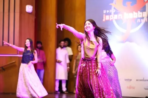 A woman in a maroon skirt dances on stage, pointing forward with a smile. Other performers in traditional attire dance behind her. The backdrop reads 'Japan Habba', suggesting a cultural event.